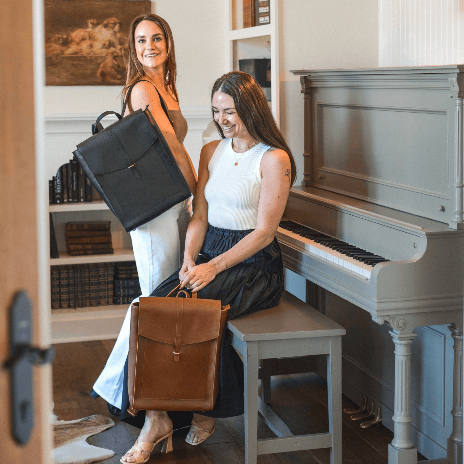 Two women holding Atlas Backpacks near a piano